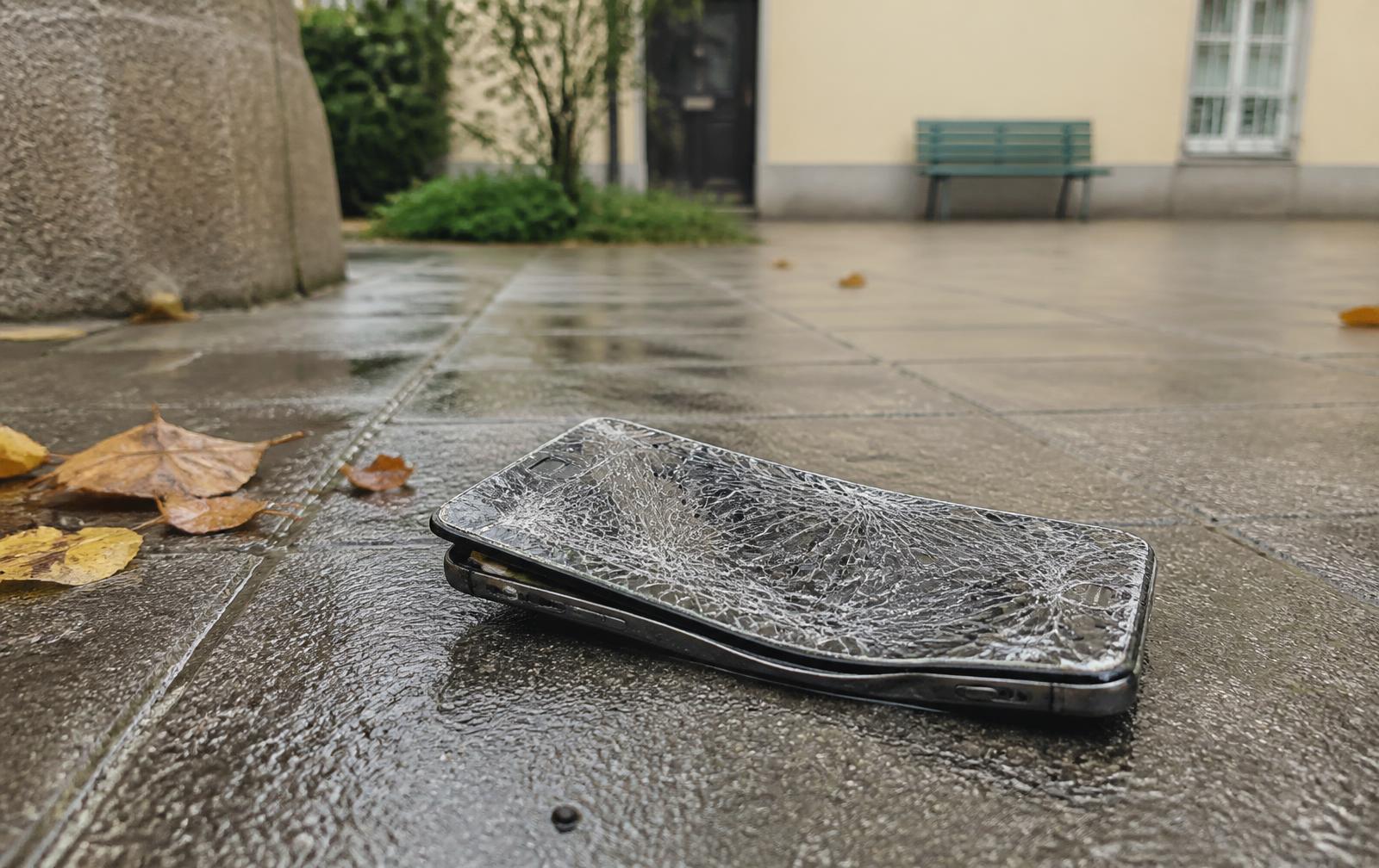Stark beschädigtes Smartphone mit zersplittertem Display auf nassem Pflaster in einem Innenhof